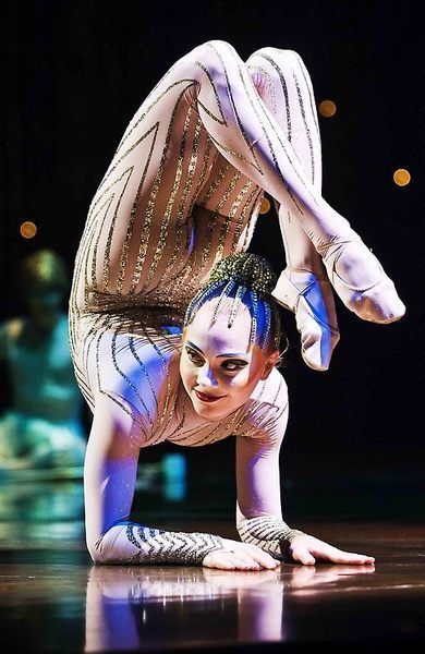 a woman in a white and gold sparkly bodysuit is contorting her body on stage, with her legs bent behind her head. she is performing a contortionist act for an audience.