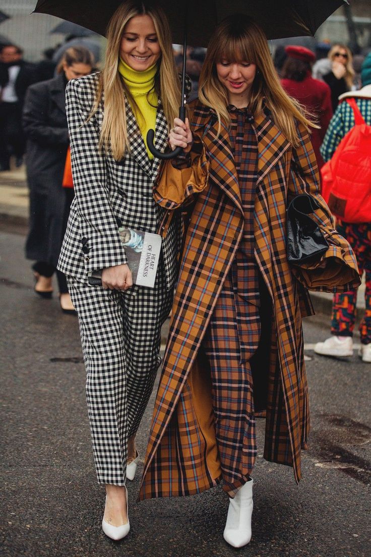 two stylishly dressed women walk down a city street on an autumn day, sharing an umbrella. one woman wears a black and white gingham pantsuit and a yellow turtleneck, while the other wears a long, brown and orange plaid coat and matching pants. both women carry handbags and are wearing white shoes. the overall image conveys a sense of elegance and sophisticated street style.