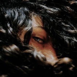 the image shows a closeup portrait of a woman with dark, curly hair partially obscuring her face. only one of her piercing blue eyes is visible, creating an air of mystery and intrigue. the lighting is lowkey, casting soft shadows on her face and adding to the overall moodiness of the image.