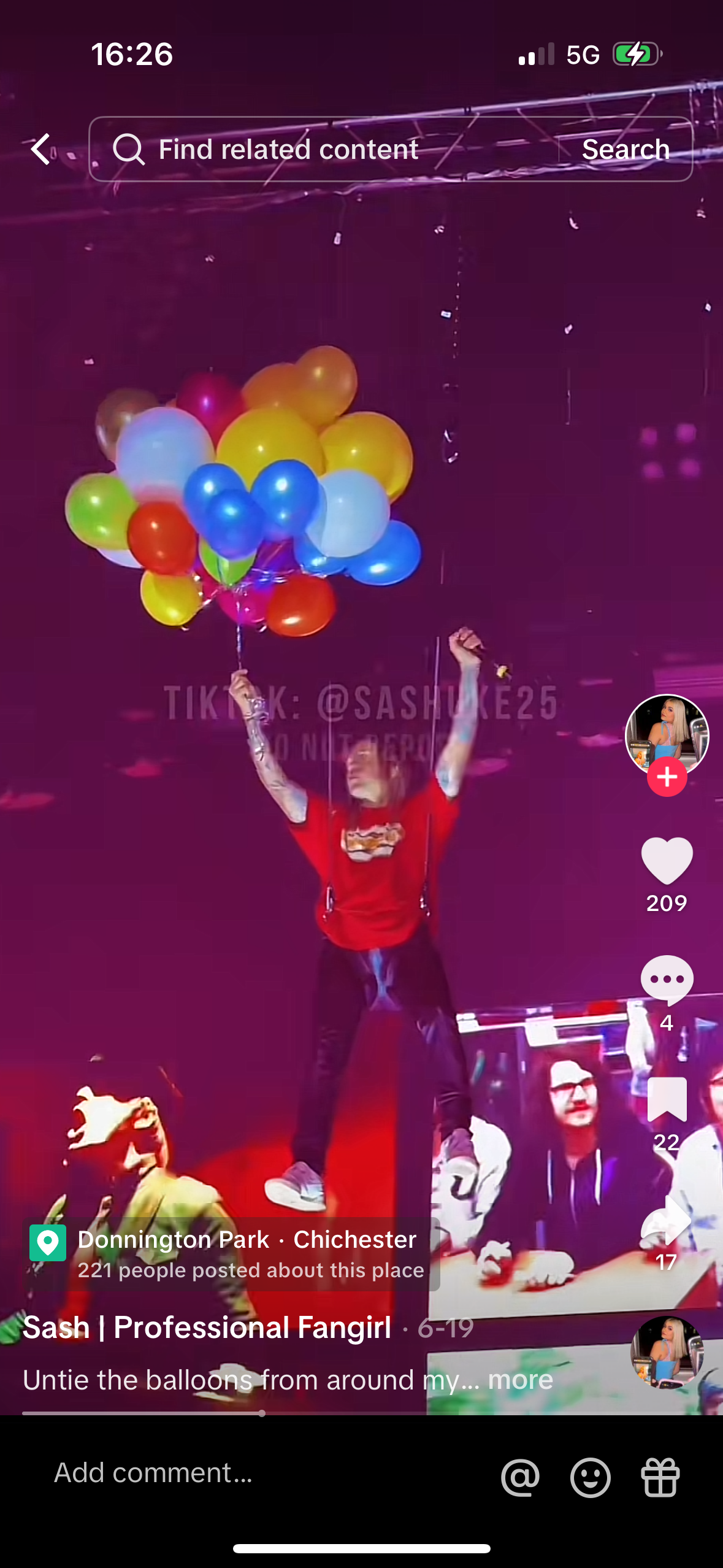 a woman is holding a bunch of balloons in the air. there are many people in the crowd and they are all having fun. the woman is wearing a red shirt and black pants.  she is standing on a stage with a microphone in hand. the woman is holding a bunch of balloons that are pink, blue, green, yellow and orange.