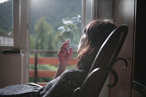 the image shows a young woman with long brown hair sitting in a darkcolored rocking chair. she is smoking a cigarette, and the smoke is visible in the air. she's positioned in front of a window that looks out onto a scenic view of mountains and trees. the overall mood is dark, contemplative, and somewhat melancholy. the lighting is soft and diffused, creating a somber atmosphere. the woman's expression is not fully visible but seems introspective. the scene suggests a moment of quiet reflection or contemplation.