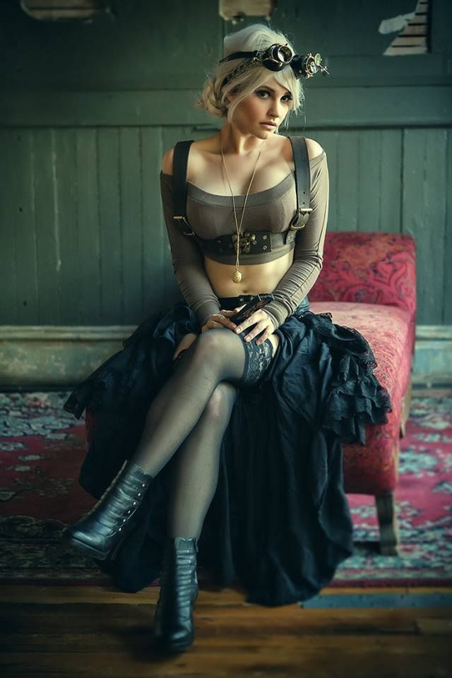 a woman in a steampunk outfit is sitting on a couch. she is wearing a brown corset, a black skirt with lace trim, black boots, and goggles. she is also wearing a silver choker and a silver necklace. her hair is blonde and she is looking at the camera with a serious expression. the room is dark and there is a red rug on the floor.