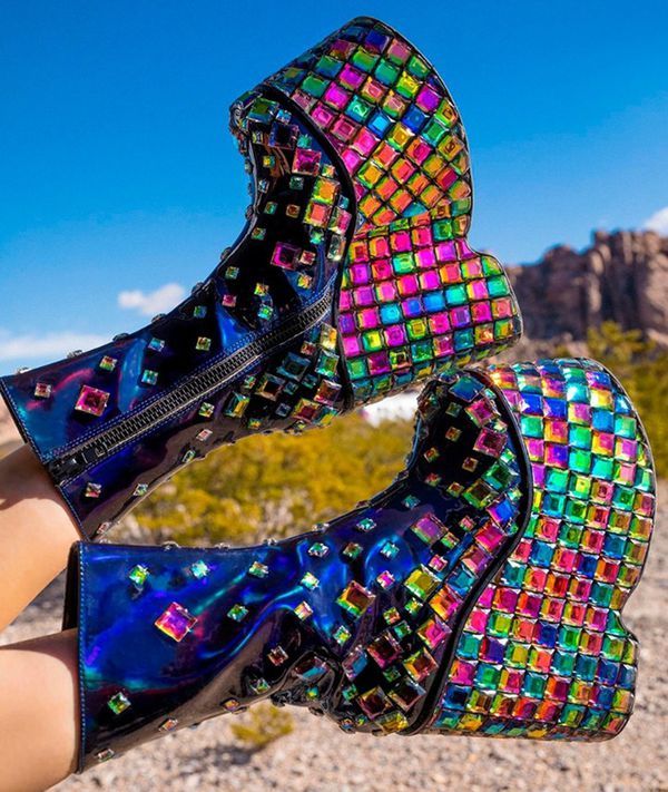 two iridescent platform boots with colorful rhinestone embellishments are held up in front of a desert landscape. the boots are blue with multicolored square rhinestones. one boot has a zipper. the background features a clear blue sky and desert vegetation.