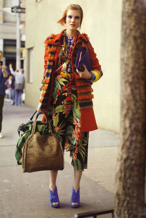 a woman is walking down a city street, she is wearing a colorful floral dress and red jacket with a fringe detailing, a pair of blue heels, and a few accessories including a brown handbag and a black clutch. she is holding the handbag in her left hand and the clutch in her right hand. the scene is shot from a slightly low angle.  she is stylishly dressed and seems to be enjoying herself.