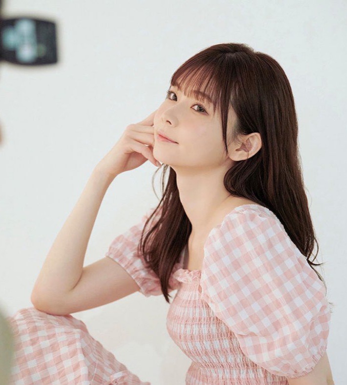 young woman with long brown hair and bangs is posing for a photoshoot. she is wearing a pink gingham dress and sitting on the floor with her hand on her chin, looking directly at the camera. she is lit by soft, natural light, giving the photo a bright and airy feel. the setting appears to be an indoor studio with a plain white background.