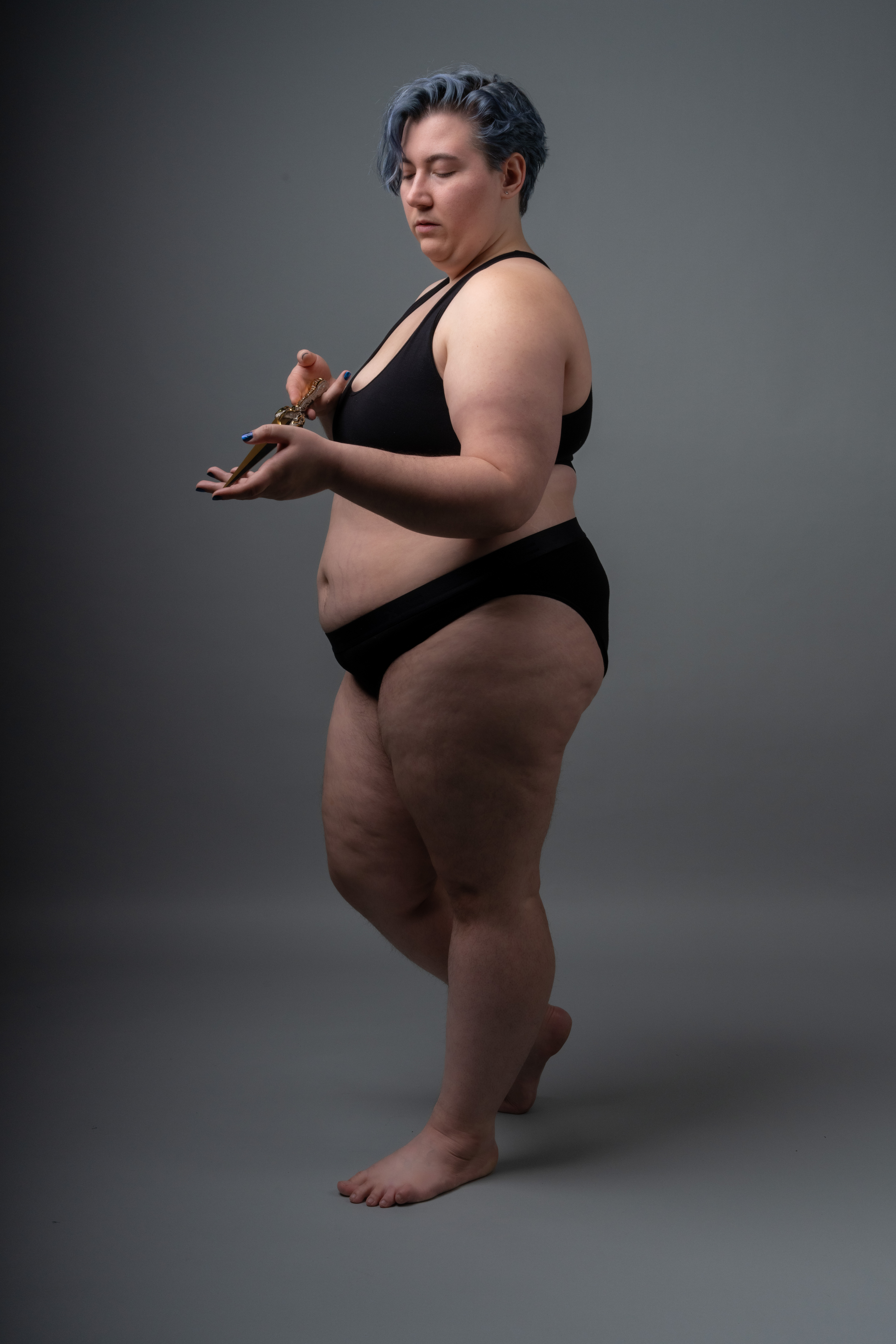 an overweight woman with blue hair stands in a studio shot, wearing black underwear and looking down at something in her hands.