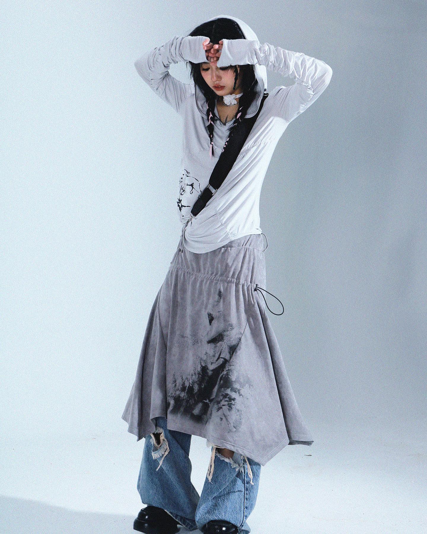 a full shot of a young woman model posing in a stylish outfit. she is wearing a light grey hoodie with long sleeves, which has a unique design and extended cuffs. the hoodie is paired with a grey, asymmetric skirt featuring a dark print, that falls over distressed light blue jeans.  she also wears a thin black choker and a single strap bag hangs across her torso. the overall style is grunge, alternative, and unique, showcasing a blend of textures and an artistic print on the skirt. the photograph is taken in a clean studio environment against a white background.