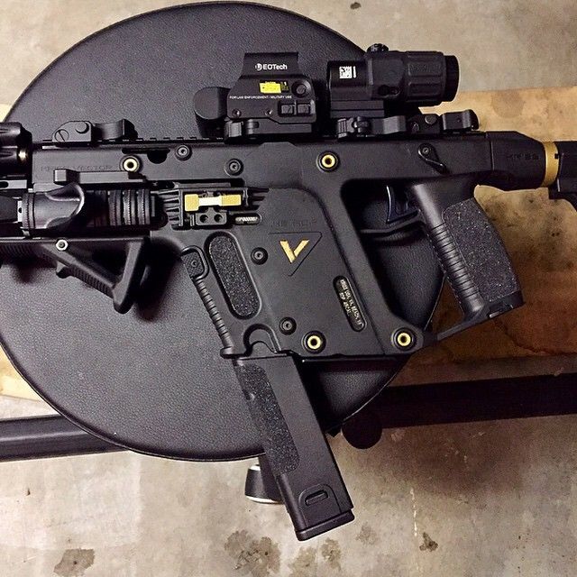 a black kriss vector submachine gun is lying on a black circular surface. the gun has gold accents and is equipped with a sight, optic, foregrip, and a magazine inserted.
