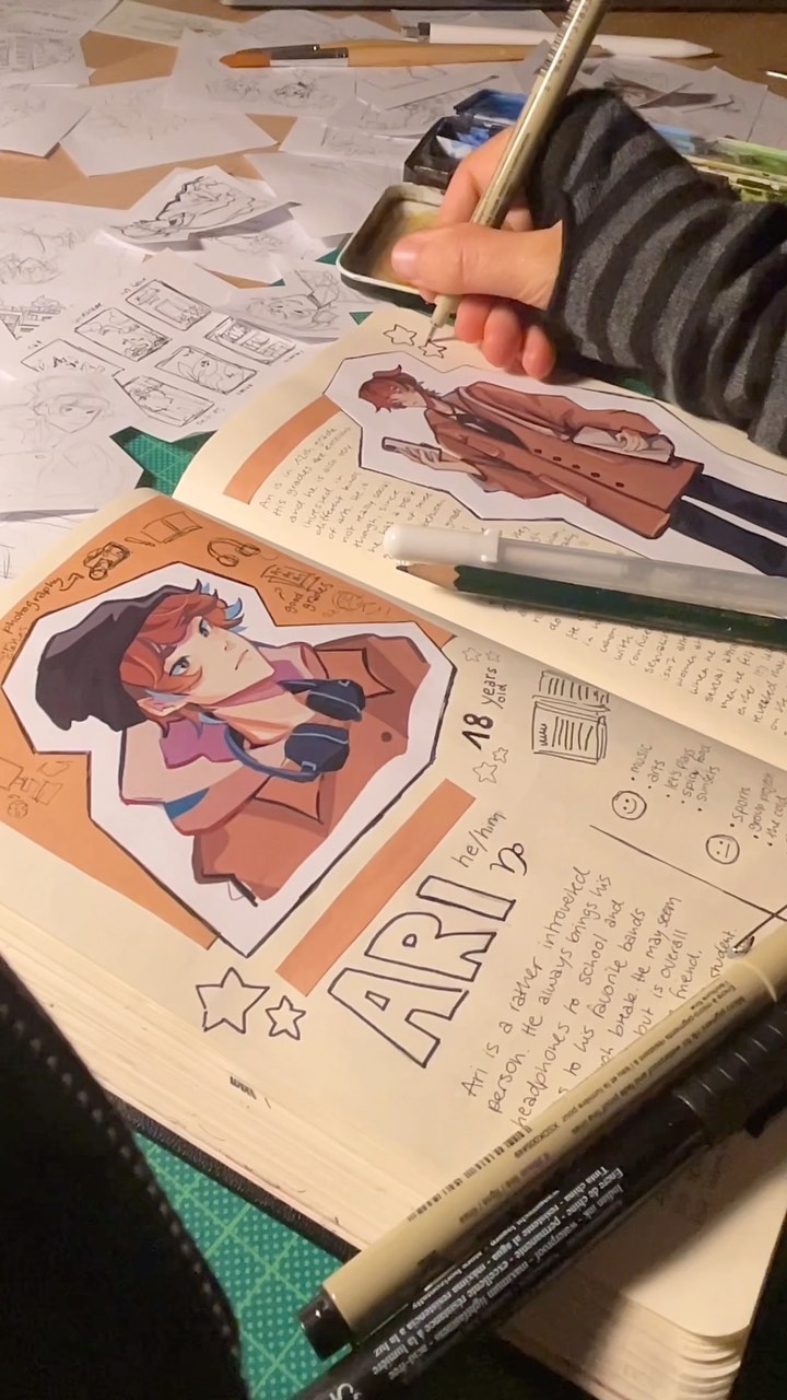 a closeup shot of an artist's workspace, focusing on an open sketchbook filled with vibrant illustrations.  the artist's hand is visible, holding a pen and working on a character design.  the sketchbook contains sketches, notes, and character profiles, suggesting a creative process in progress.  various art supplies, including pens, markers, colored pencils, and a watercolor palette, are scattered around the workspace, indicating an active and creative environment. the style of the artwork is reminiscent of anime or manga.  the overall scene is busy and suggests a creative process where the artist is actively working on their art.