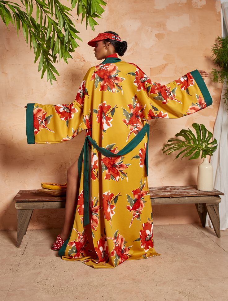 a back view of a woman wearing a long, mustard yellow kimono adorned with a large red floral print. the kimono has long sleeves and a green trim along the edges. the model also wears a red visor and red shoes. she is standing in front of a rustic wooden bench with tropical plants in the background. the overall setting and the model's attire have a relaxed and fashionable vibe.