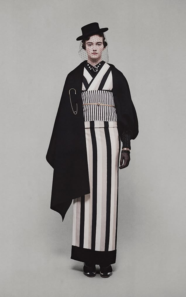 a woman stands facing forward, wearing a unique and stylish outfit. she has a black hat with a veil and is wearing a black shawl over a striped kimonostyle dress with a striped obi belt. a large silver safety pin is attached to the shawl. she also has black gloves. the background is a plain, light gray color.