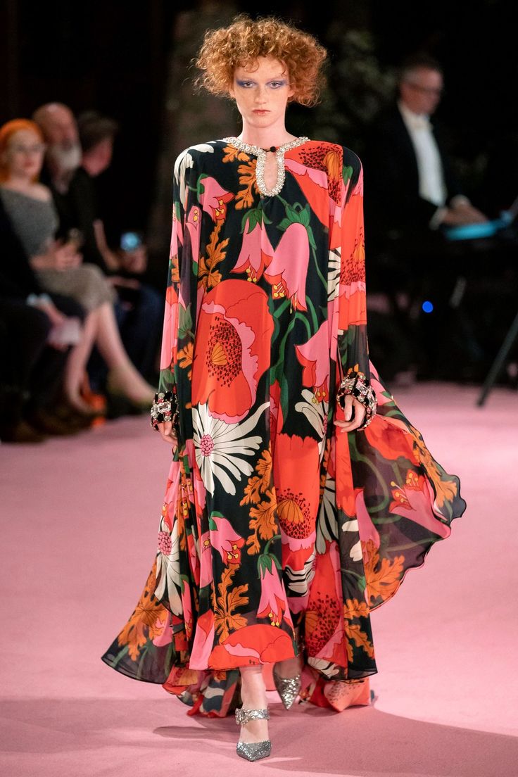 a model with curly red hair walks the runway wearing a long, flowing maxi dress with a bold floral print featuring poppies and other flowers in shades of red, pink, orange, and black on a dark background.  the dress has long sleeves and appears to be made from a light, flowing fabric. the model is wearing silver heels and the background shows other people in the audience, somewhat blurred. the overall aesthetic is reminiscent of 70s fashion. the image appears to be taken at a professional fashion show.