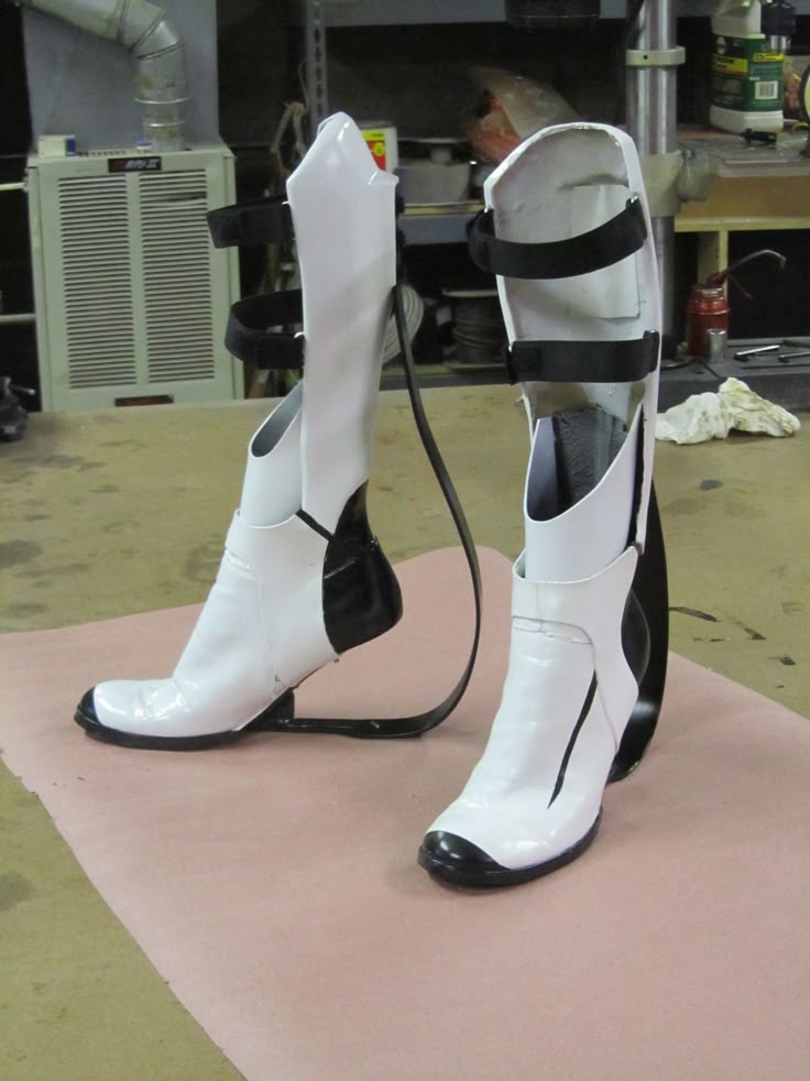 two futuristic white and black boots with straps are standing on a surface covered with pink cloth. the boots appear to be part of a costume, possibly cosplay. they have a unique design with black accents and straps, and they are positioned against a background with various objects on shelves and a gray ventilation unit.