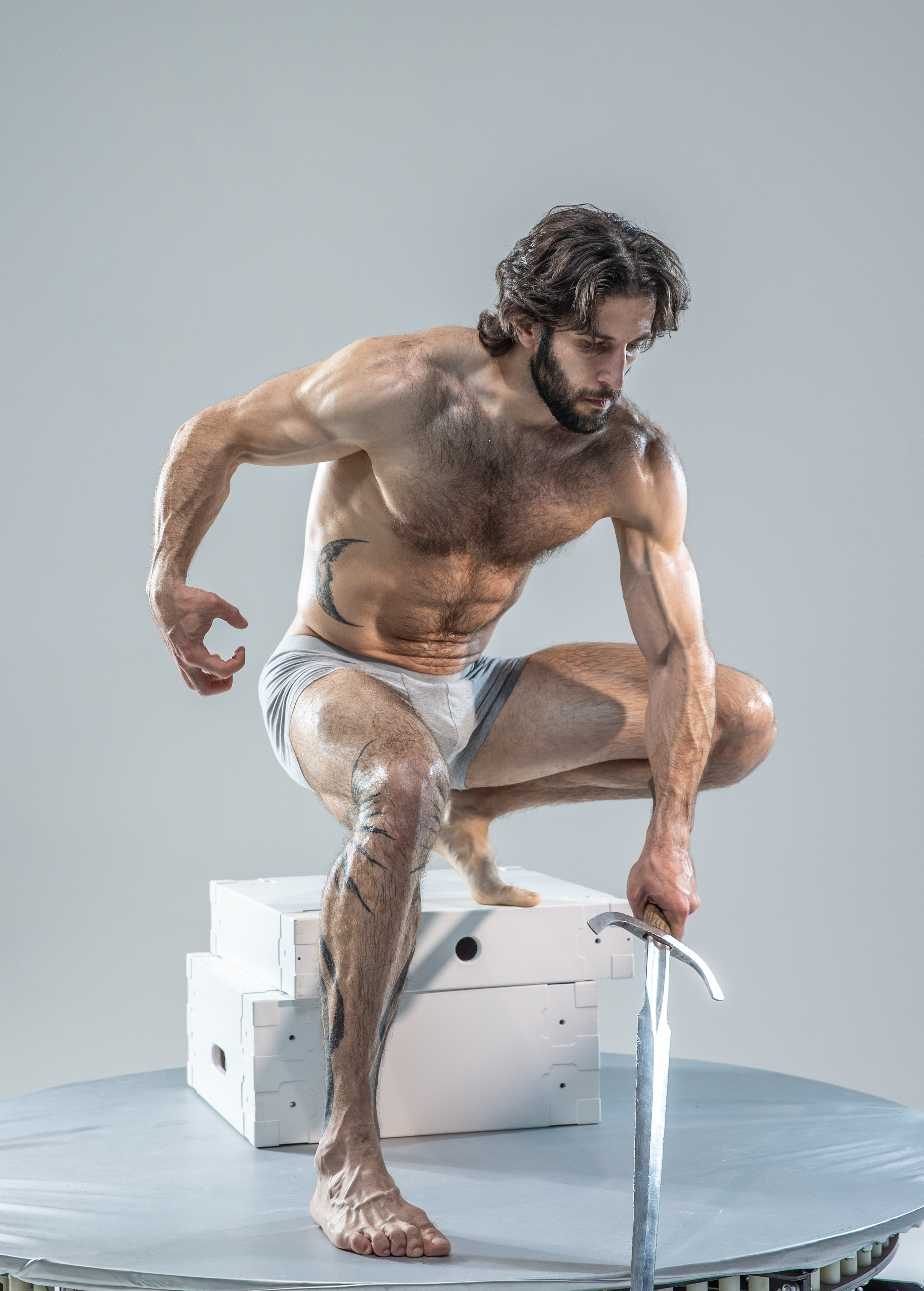 a muscular man with long, dark hair stands in a dynamic pose on top of two white cubes. he is shirtless, showcasing his physique, and wears only white underwear. he grips a silver sword in his right hand, the tip pointed towards the ground. the man's gaze is directed downwards, focused on the sword. the background is a neutral gray, illuminated with soft studio lighting. the overall image evokes a sense of power, mythology, and strength.