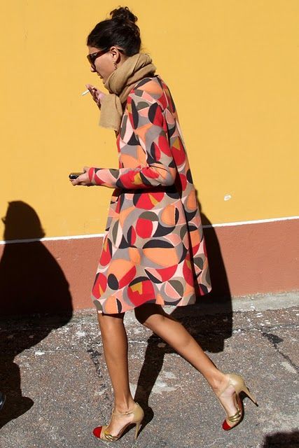 a stylish woman in a geometric print coat, sunglasses, and a beige scarf is walking down a yellowwalled street. she is smoking a cigarette and wearing gold and red high heels. her shadow is cast on the ground beside her. the overall aesthetic is chic and autumnal.