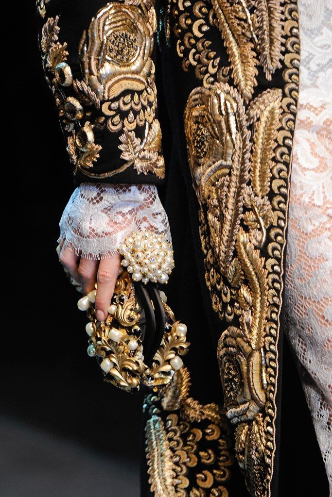 loseup detail shot of a dolce & gabbana garment showcasing intricate gold embroidery on black fabric. the image focuses on the sleeve and a portion of the bodice, highlighting the rich texture and opulent design.  a hand adorned with a pearl and gold bracelet holds a similarly ornate clutch. the detail shows the high level of craftsmanship.  the model is wearing delicate lace underneath the jacket.