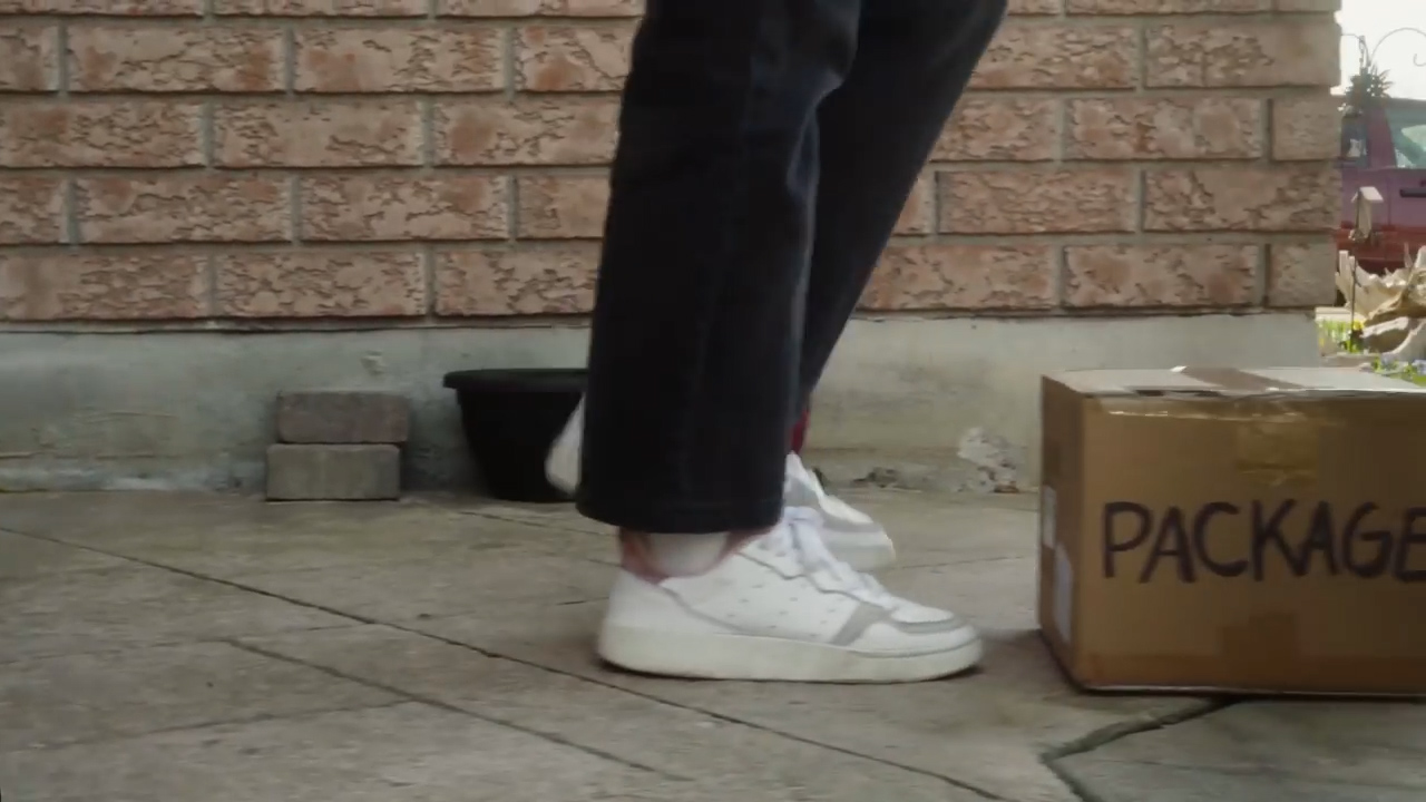 a person wearing black pants and white sneakers is standing on a concrete step in front of a brick wall. they are about to pick up a cardboard box labeled "package" that is sitting on the step. the image is taken from a high angle, and the lighting is natural daylight.
