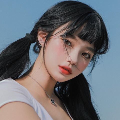 a closeup shot of a young woman with black hair styled in pigtails and blunt bangs. she has fair skin, rosy cheeks, and is wearing red lipstick. she is wearing a white top and a silver necklace. an earring is visible in her left ear. the background is a clear blue sky.