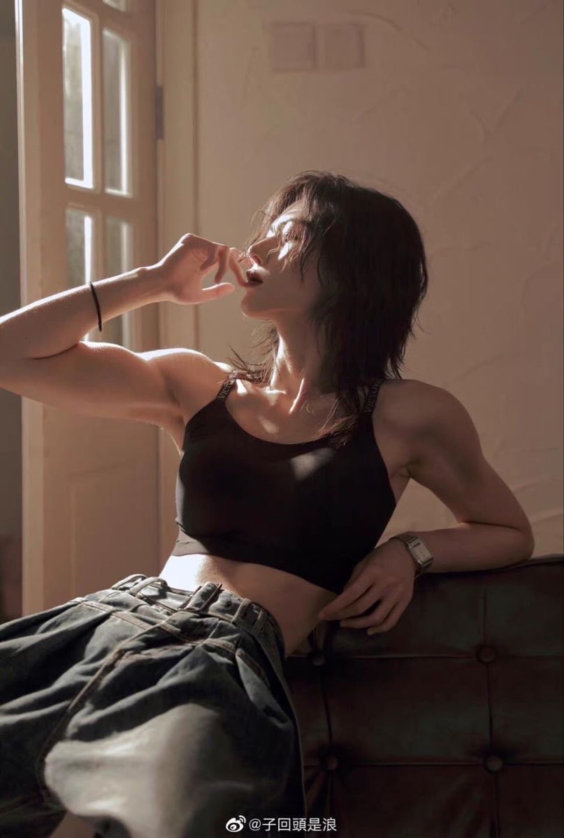 the image shows a woman with short dark hair, wearing a black sports bra and a denim skirt. she is leaning back against a brown leather couch, with one arm raised and her hand touching her face. her other arm is resting on the couch, and she is wearing a watch on her wrist. the sunlight is streaming in through a window behind her, casting warm light on her face and upper body. the background is blurred, drawing attention to the woman in the foreground.