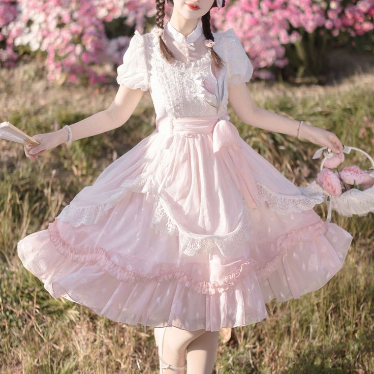 a young woman wearing a pink lolita fashion dress is standing in a garden. the dress has a white lace overlay and a pink bow at the waist. she is holding a hand fan in her right hand and a basket of pink flowers in her left hand. the background is a blur of pink and white flowers.