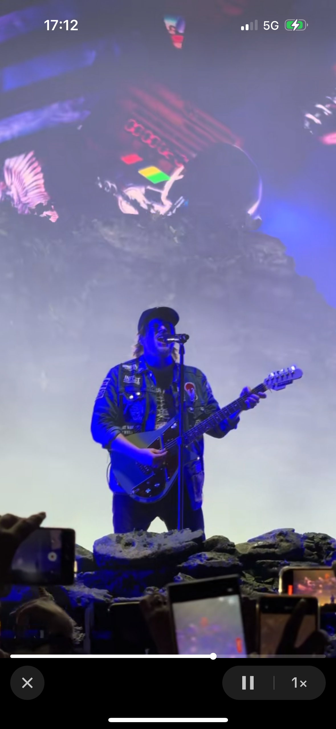 a musician is performing on stage at a concert.  he is holding an electric guitar and singing into a microphone. he has long hair and a beard and is wearing a cap.  the stage is lit with blue and white lights.  the audience is not visible, but their cell phone screens can be seen.