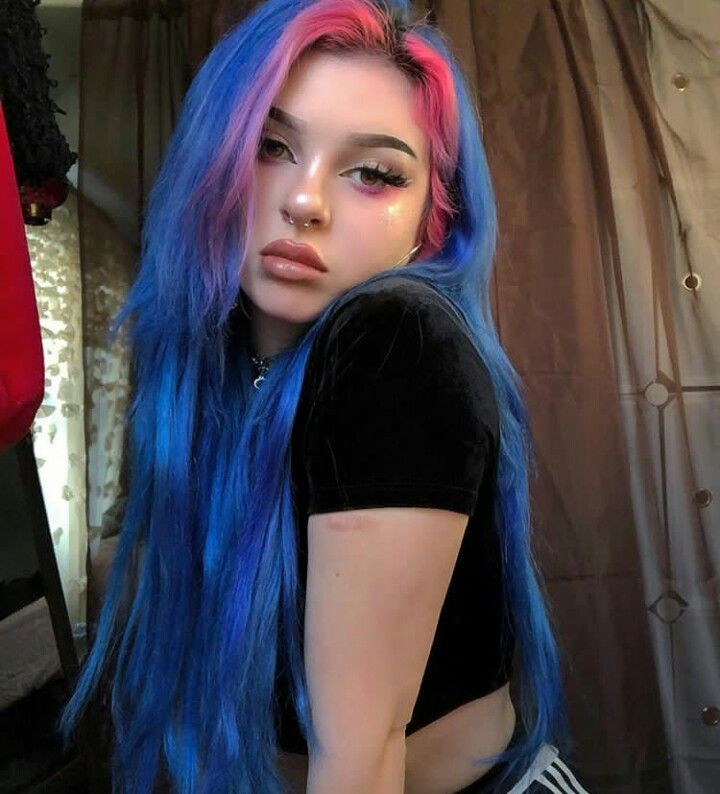 a young woman with blue and pink hair poses for a portrait. she wears a black crop top and has elaborate makeup on, including glitter and dramatic eyeliner. a nose ring and necklace adorn her face and neck. the background is a curtain and glimpses of other objects.