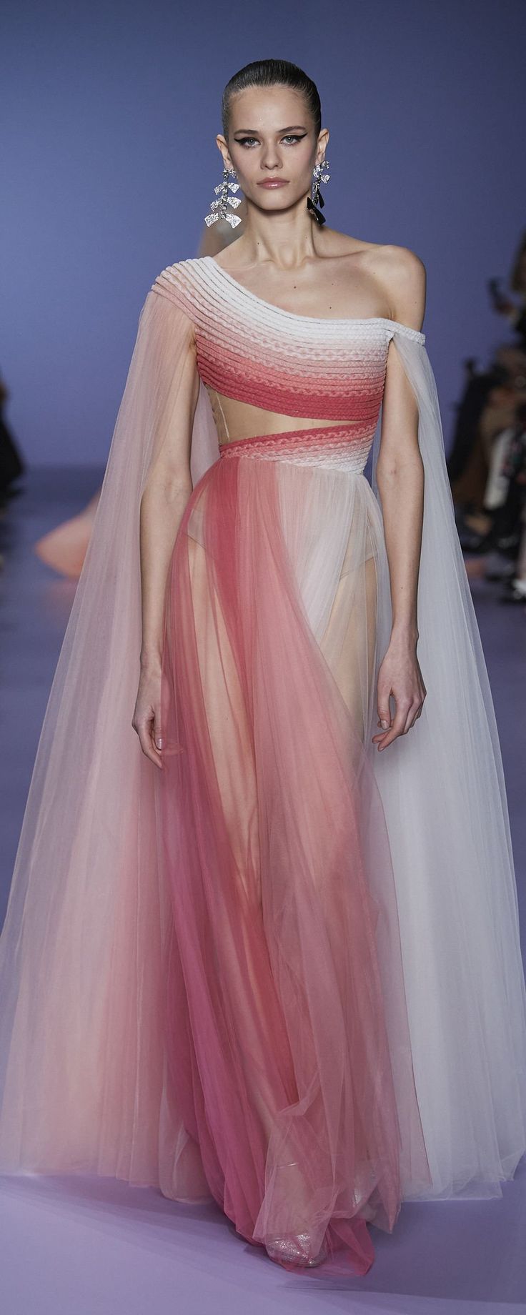 a model walks the runway wearing a stunning ombre gown. the dress features a oneshoulder design with a sheer tulle overlay in shades of pink, red and white. the gown has a capelike overlay that drapes elegantly from the shoulders. the model is wearing delicate earrings and minimal makeup, allowing the dress to be the focal point of the image.  the background suggests a fashion show setting.