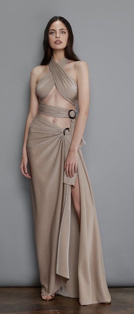 a full shot of a female model posing in a long, beige metallic dress.  the dress is a halter style that wraps around the body, with cutouts at the waist and a high slit on one side. the model has long brown hair and neutral makeup. she is standing on a gray background, and the floor is visible at the bottom of the image. the overall style is elegant and sophisticated.