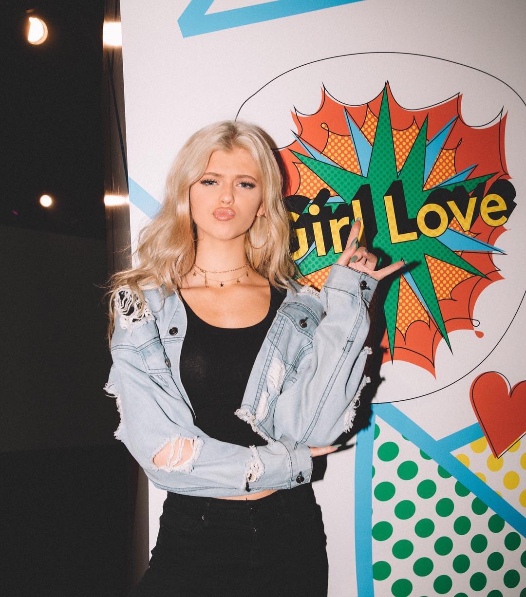 a young woman with blonde wavy hair is posing in front of a pop artstyle background that says "girl love". she is wearing a black tank top, black jeans, and a ripped denim jacket. she has multiple necklaces on. her lips are pursed in a pout, and she's making a hand gesture with her right hand. the background features bright colors, polka dots, and a red heart. the image is taken indoors.