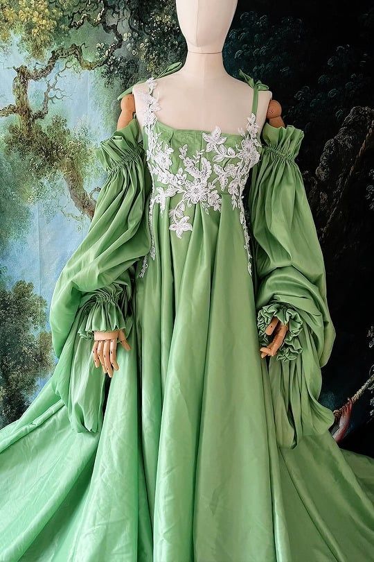 a green satin gown with lace detailing, puffy sleeves, and an offtheshoulder neckline is displayed on a mannequin. the dress has a long, flowing skirt. the mannequin is against a background that looks like a painted mural of a forest scene.