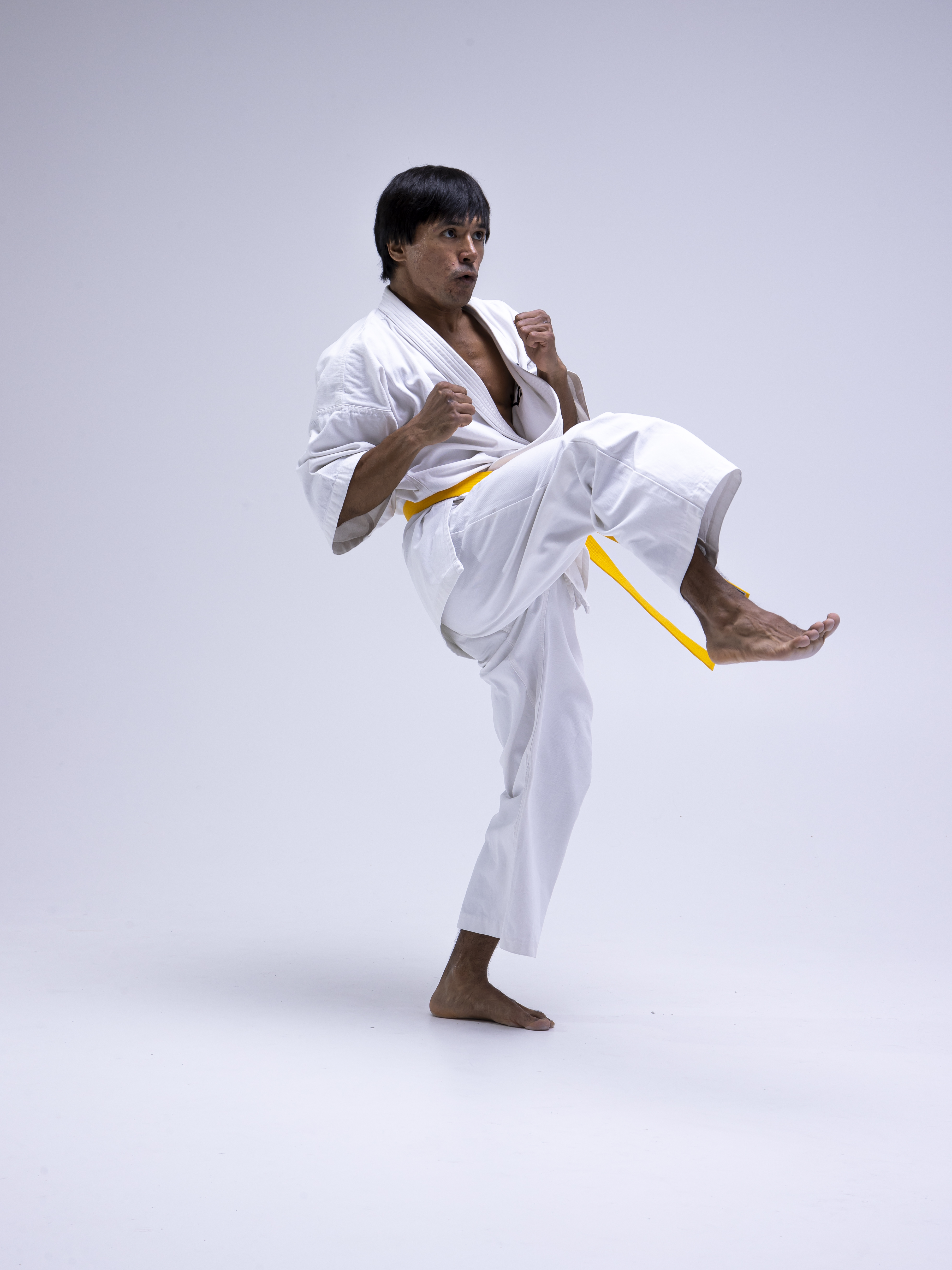 a man in a white karate uniform with a yellow belt is performing a kick. he is barefoot and indoors against a plain white backdrop. he is in a martial arts pose, ready to fight.