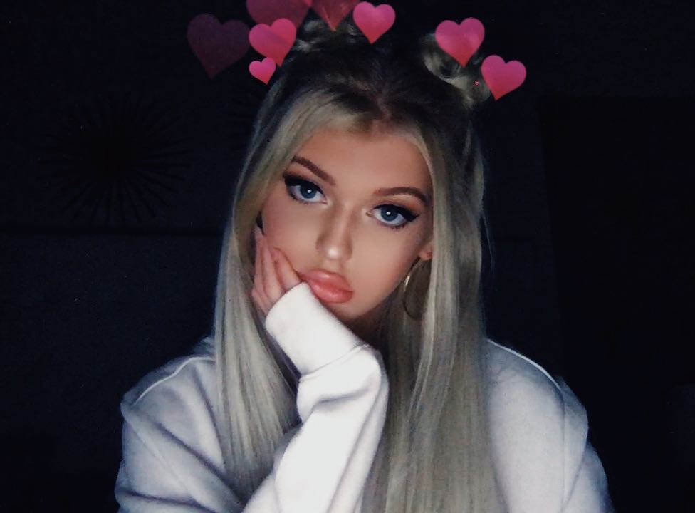 a photo of a young woman with blonde hair and blue eyes, wearing a white hoodie and a filter that adds pink hearts above her head. she has makeup on and is pouting with her hand resting on her face. the background is dark.