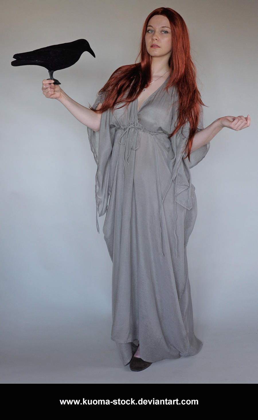 a fullbody shot of a young woman with long, flowing red hair.  she's wearing a flowing, light grey maxi dress that has a loose, almost medieval or fantasyinspired style.  the dress is cinched at the waist with a drawstring. she stands calmly, holding a black ravenlike bird in one hand, her other hand outstretched slightly. the backdrop is a plain light grey, focusing attention on the model and her attire. the overall impression is one of mystery and a slightly mystical or magical aesthetic. the watermark www.kuomastock.deviantart.com is visible at the bottom.