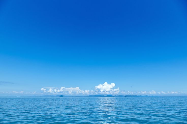 the image shows a calm ocean under a clear blue sky with scattered clouds on the horizon. the water is blue and appears still with faint ripples, suggesting a peaceful day. in the distance, a coastline is visible with small landforms that could be islands or headlands.
