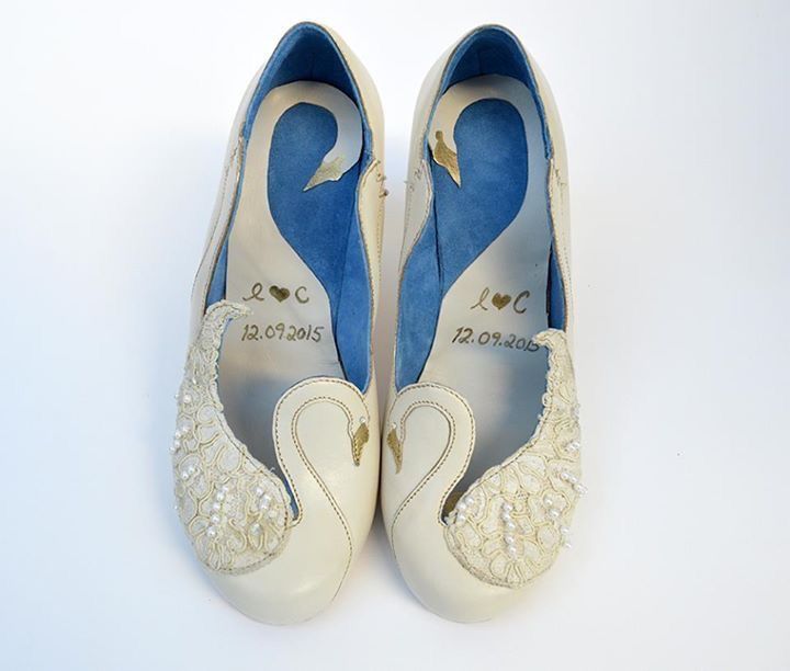 a pair of ivorycolored bridal shoes is displayed on a white background. the shoes feature a swan design on the toe, with the swan's body formed by the shoe's upper and its head curving towards the heel. the swan's body is adorned with lace and pearl embellishments. the inner lining of the shoes is blue. inside each shoe, there's a monogram of the letters "l" and "c" with a heart between them, along with the date "12.09.2015" inscribed below.
