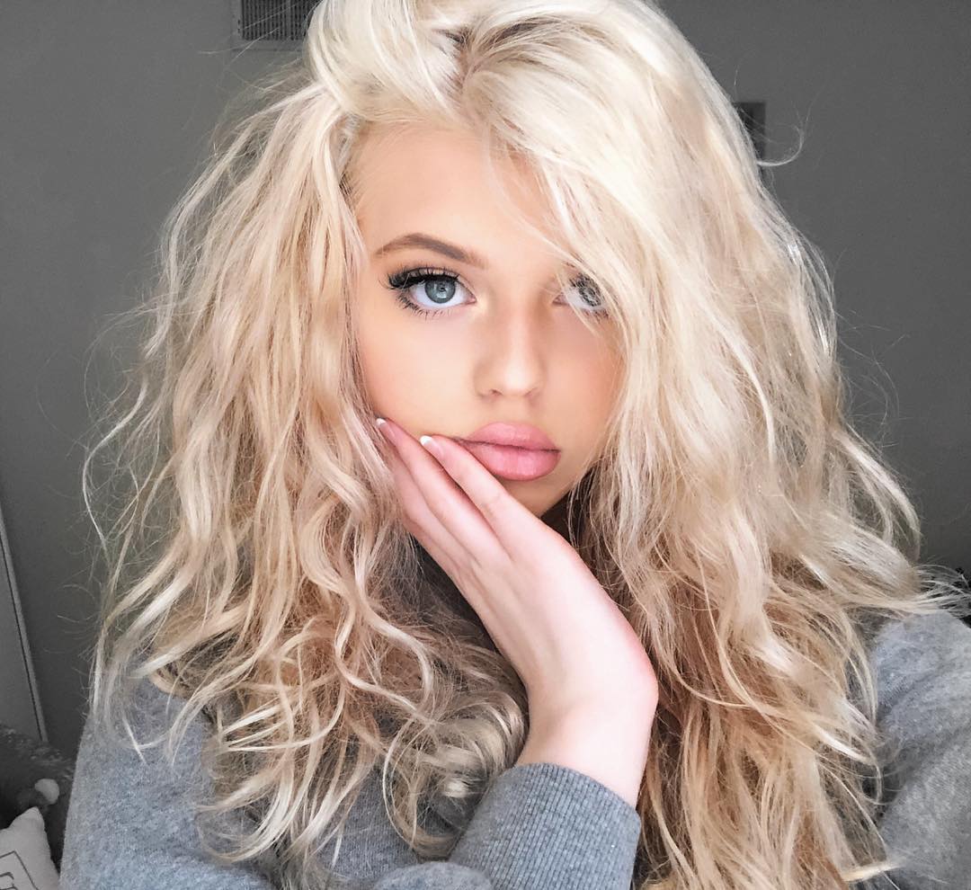 a young woman with blonde, wavy hair and blue eyes is posing for a selfie. she is wearing a gray sweater and has her hand resting on her face. she is wearing makeup, including lipstick and eyelashes. the background is a plain gray.