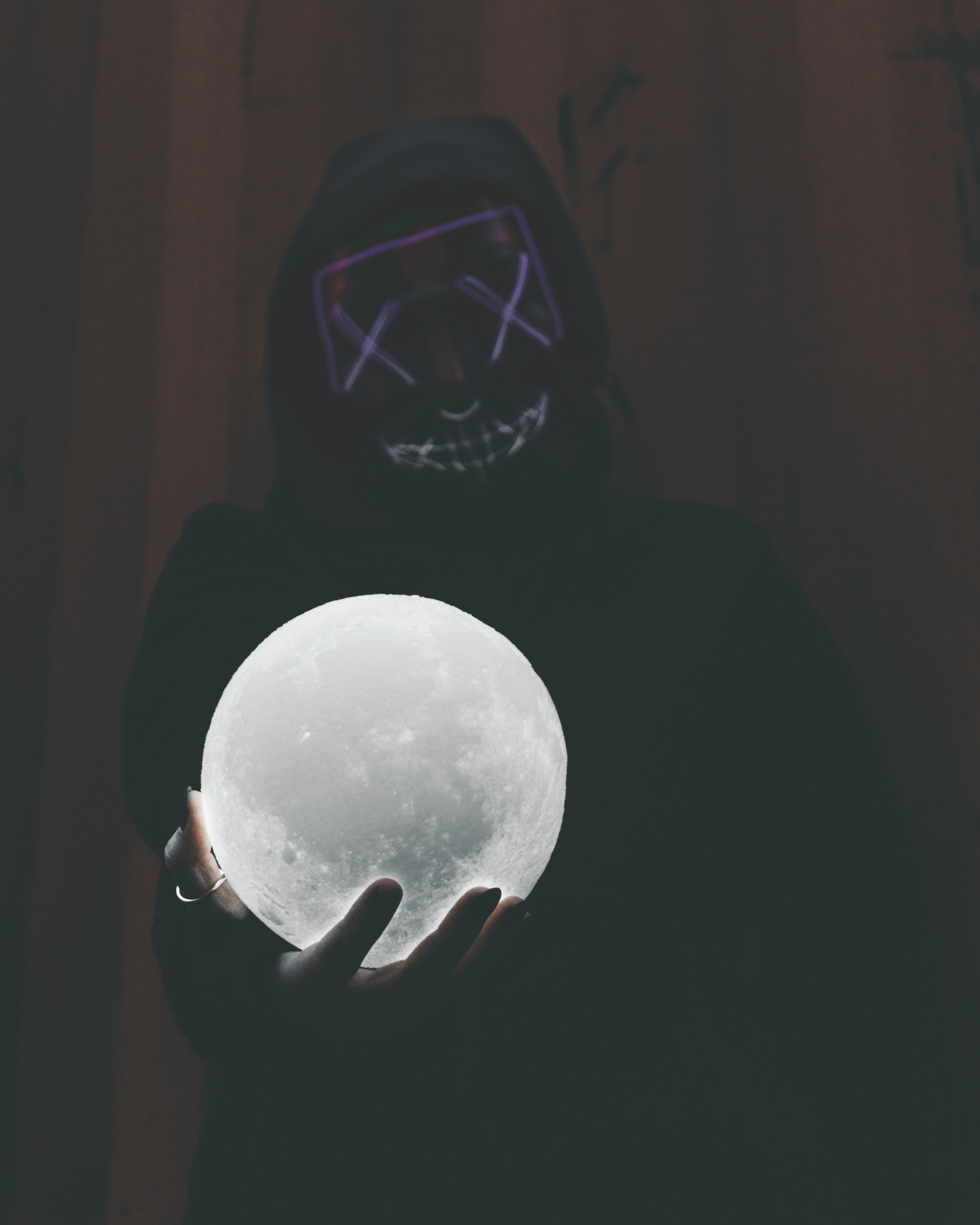 a person wearing a black hoodie and a glowing purple mask holds a glowing moonshaped object in their hands. the background is dark, and the only light source appears to be coming from the mask and the moon object.