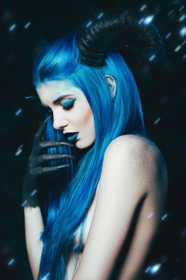 a portrait of a beautiful woman with blue hair and dark makeup, wearing a black horn and gloves. the photo was taken in a dark studio with a dark background.
