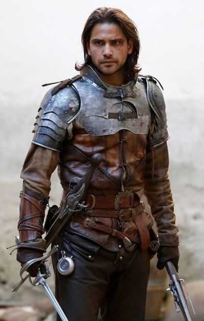 a man in medieval warrior attire stands facing the camera, holding a sword in each hand. he is clad in a combination of leather and metal armor, with intricate detailing on his breastplate and gauntlets. his expression is serious and determined. the setting appears to be outdoors, with a muted background that emphasizes the warrior's presence.