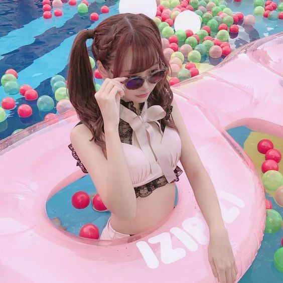 young woman with brown hair in pigtails is sitting inside a pink inflatable ring in a pool filled with colorful plastic balls. she is wearing a pink and black bikini top, sunglasses, and a smile. the photo is taken from a high angle, and the bright, natural lighting suggests a sunny day.