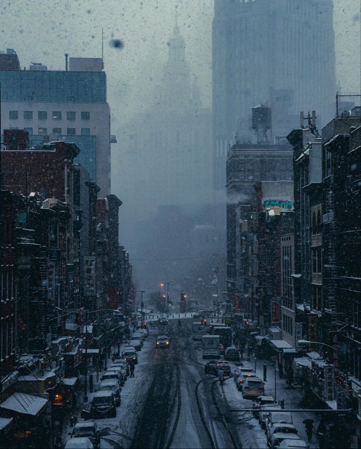 the image shows a city street blanketed in snow during a blizzard. the buildings lining the street are tall and mostly dark, creating a moody atmosphere.  the snow is falling heavily, reducing visibility.  cars are visible driving down the snowcovered street, their headlights barely piercing the thick snow.  the overall color palette is muted greys and blues, enhancing the cold and bleak ambiance. a tall building looms in the background, partially obscured by snow and mist.  the scene evokes a feeling of isolation and quiet intensity within the heart of a busy city.