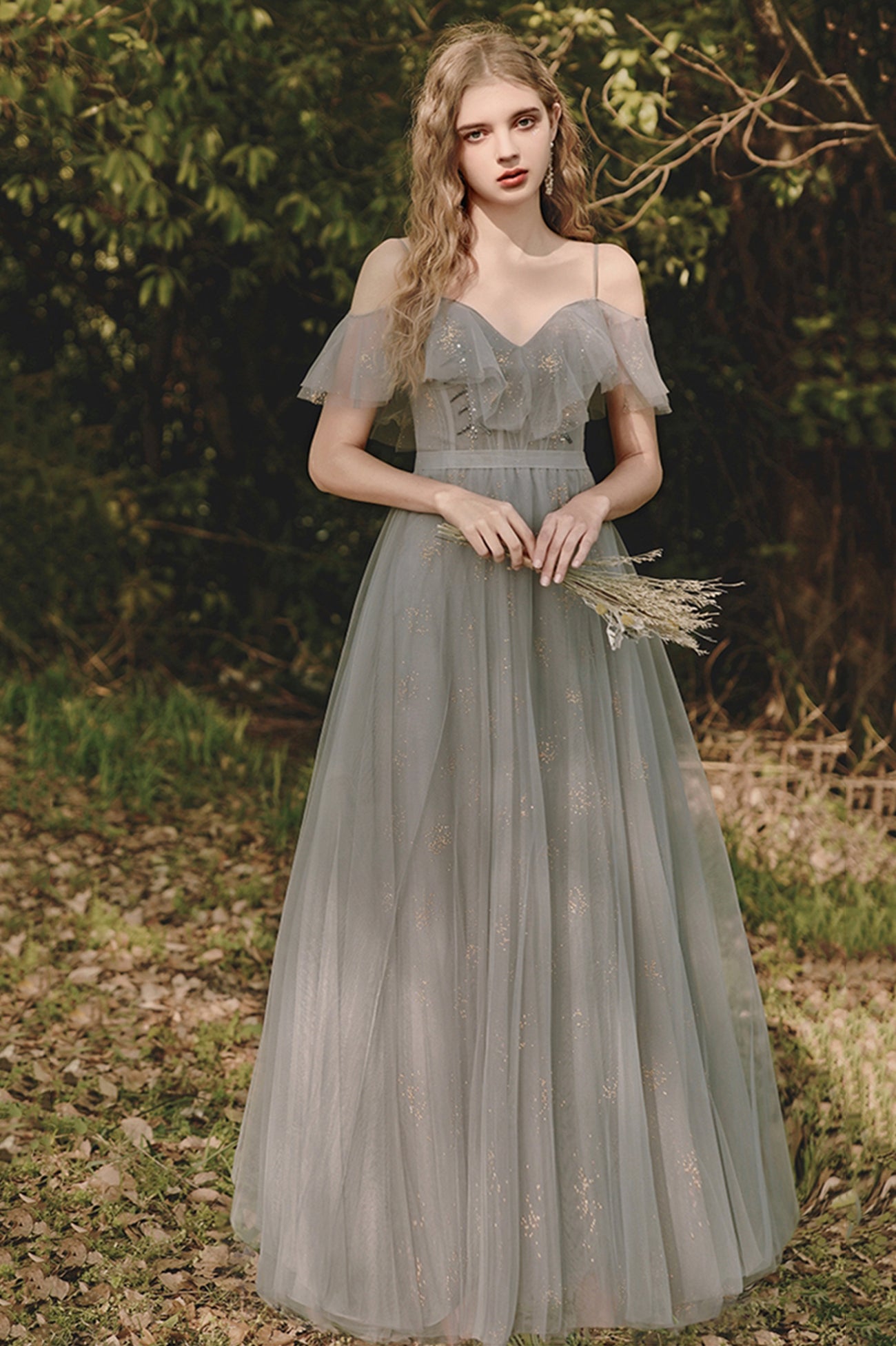 a young woman stands in a forest setting, wearing a long, graygreen tulle dress with offtheshoulder ruffled sleeves. she is holding a small bunch of dried flowers in her hands. the dress is a formal gown, likely a bridesmaid dress.