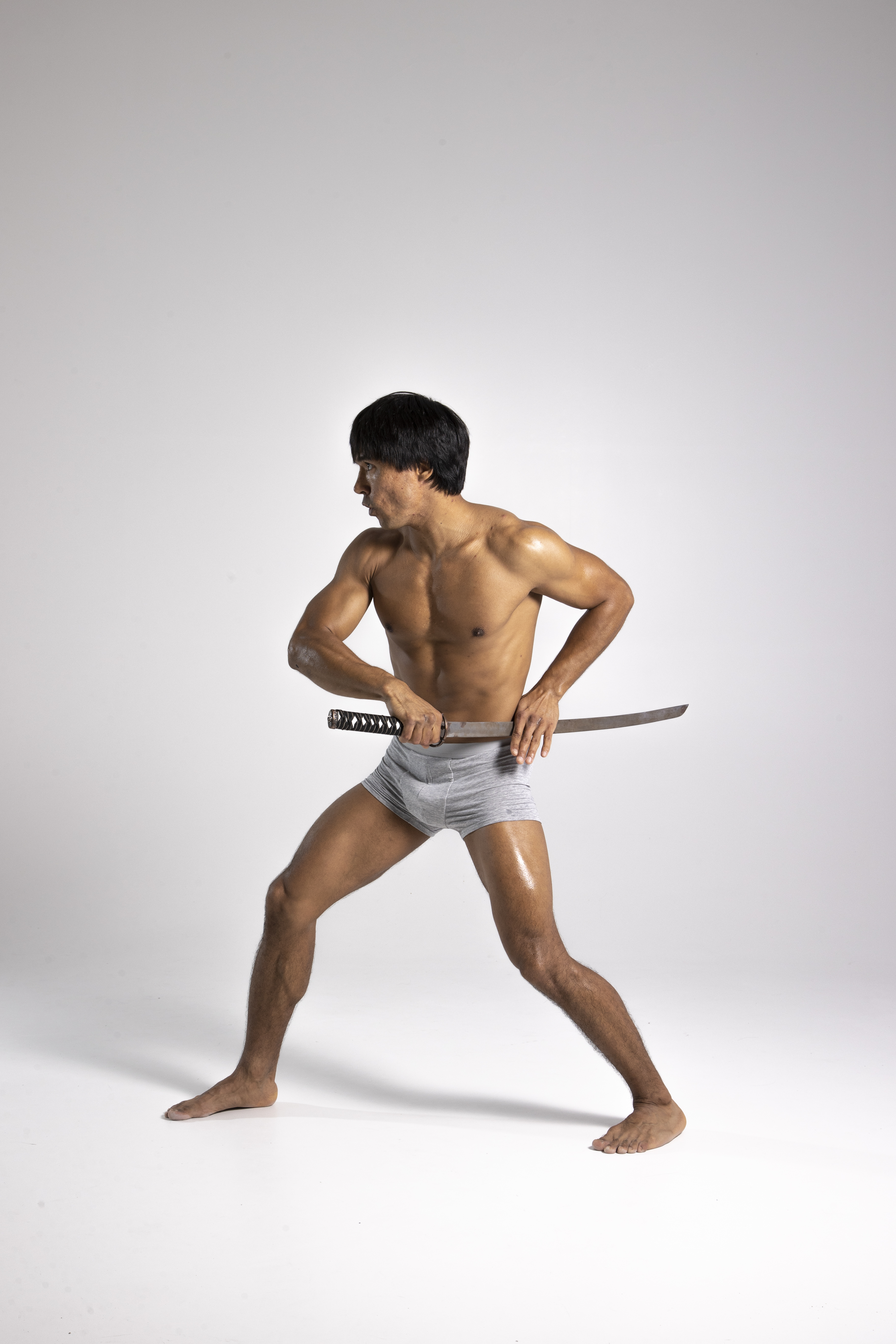 a full shot captures a muscular man, shirtless and wearing gray shorts, holding a katana while looking away in a studio setting.