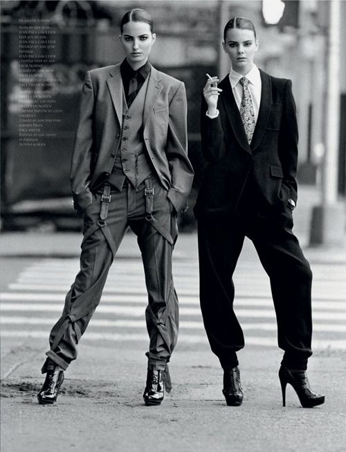 two women stand on a city street wearing suits and heels. one woman is wearing a grey suit with suspenders and a black shirt and tie. the other woman is wearing a black suit with a patterned tie and is holding a cigarette.