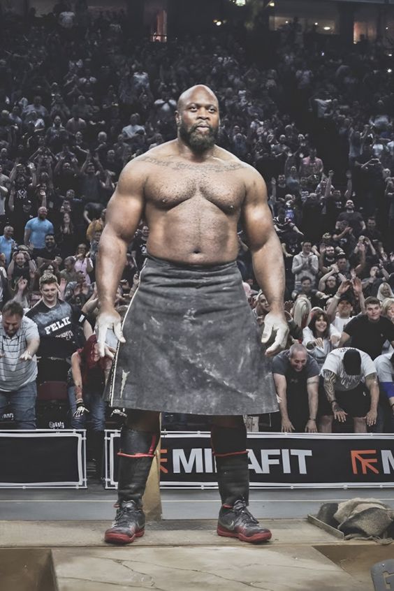 a shirtless, muscular man with a shaved head and goatee is standing on a platform in front of a large crowd in an arena. he is wearing a kilt, wrist wraps, and athletic shoes. he appears to be a strongman competitor. the audience is cheering and clapping in the background.  the image is taken from a low perspective, making the man appear larger than life.