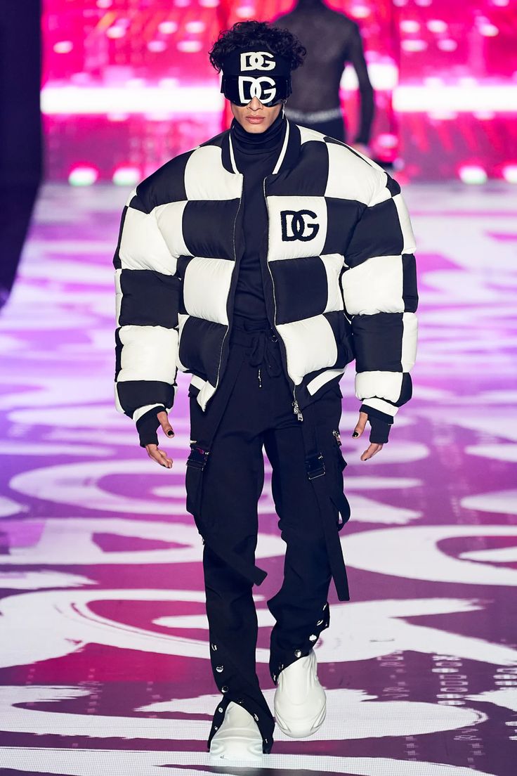 a male model walks down the runway wearing a black and white checkered puffer jacket, black pants with buckles, white shoes, and sunglasses. he is wearing a black headband with the letters dg on it.