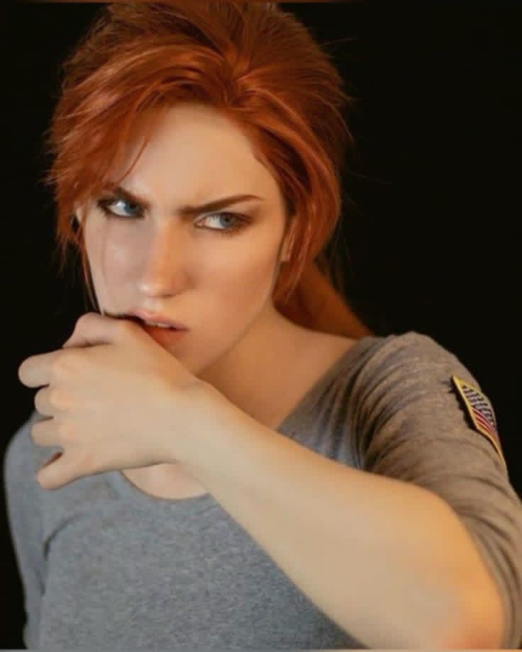 a woman with red hair is biting her fingernail and looking off to the side with a concerned expression. she is wearing a gray shirt with an american flag patch on the sleeve. the background is a dark color.