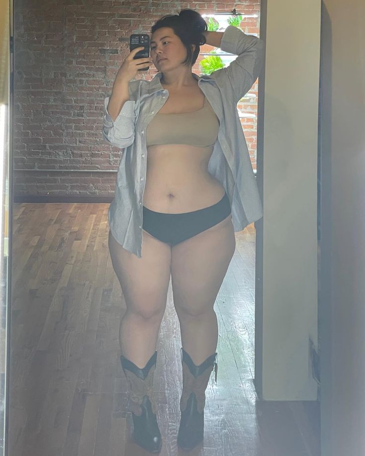 a woman is taking a selfie in a mirror. she is wearing lingerie consisting of a nudecolored top and darkcolored underwear. she is also wearing a lightcolored buttondown shirt that is open and cowboy boots. her hair is in a bun. the background shows a brick wall and a wooden floor. the image promotes body positivity.