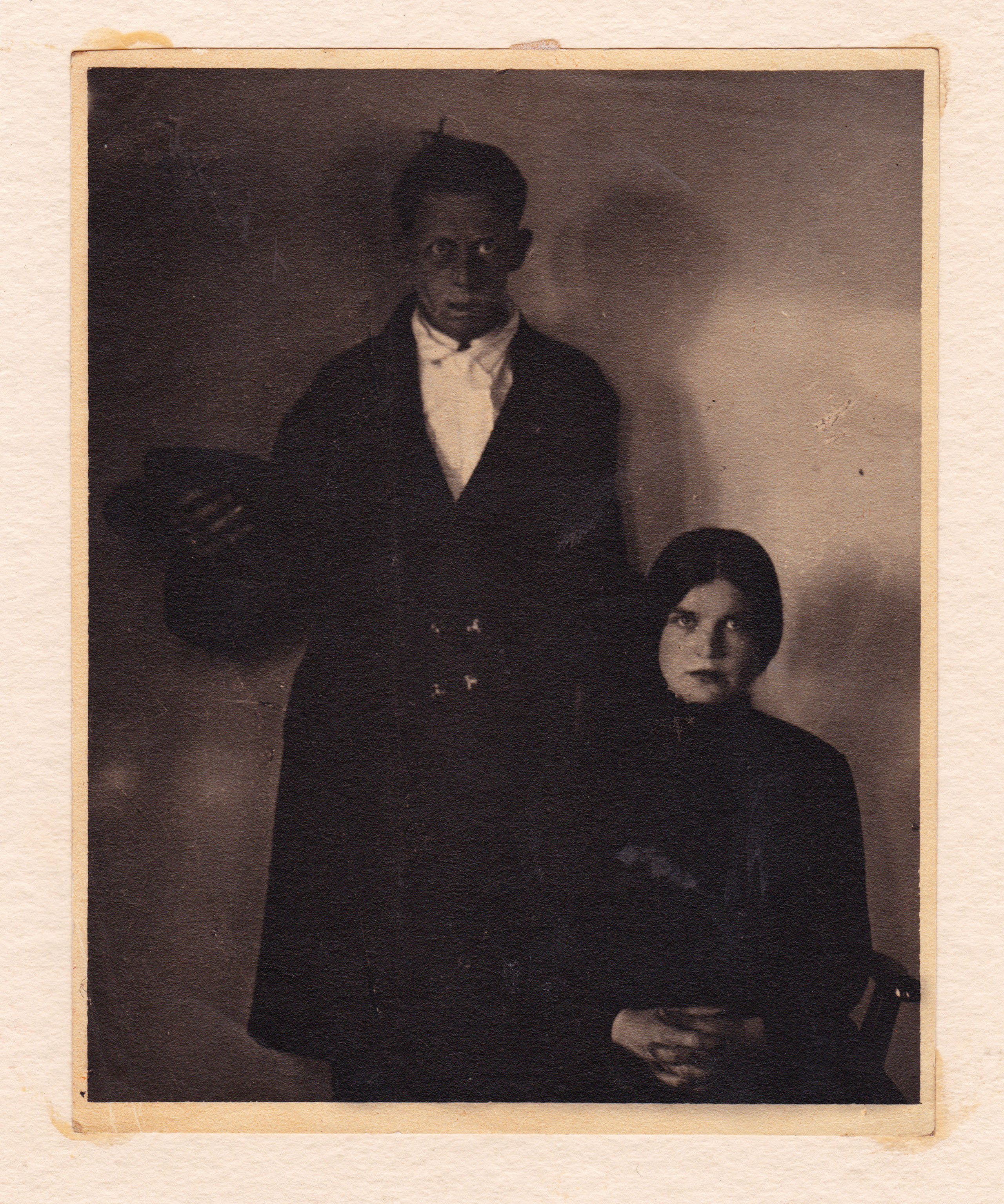the image is a vintage black and white photograph, possibly sepiatoned, depicting a couple posed formally for a portrait. the man, appearing to be of african american descent, is dressed in a dark suit with a lightcolored collared shirt and what appears to be a bow tie. he holds a hat in his hand.  the woman, whose race is less distinct in the photo's contrast but is likely caucasian, wears a darkcolored dress with a high neckline.  both individuals have serious expressions. the background is a plain, lightcolored wall, typical of studio portraits from the early to mid20th century. the overall style and clothing suggest the photo was likely taken sometime in the 1920s.  the image is presented within the borders of a slightly aged photograph mount.