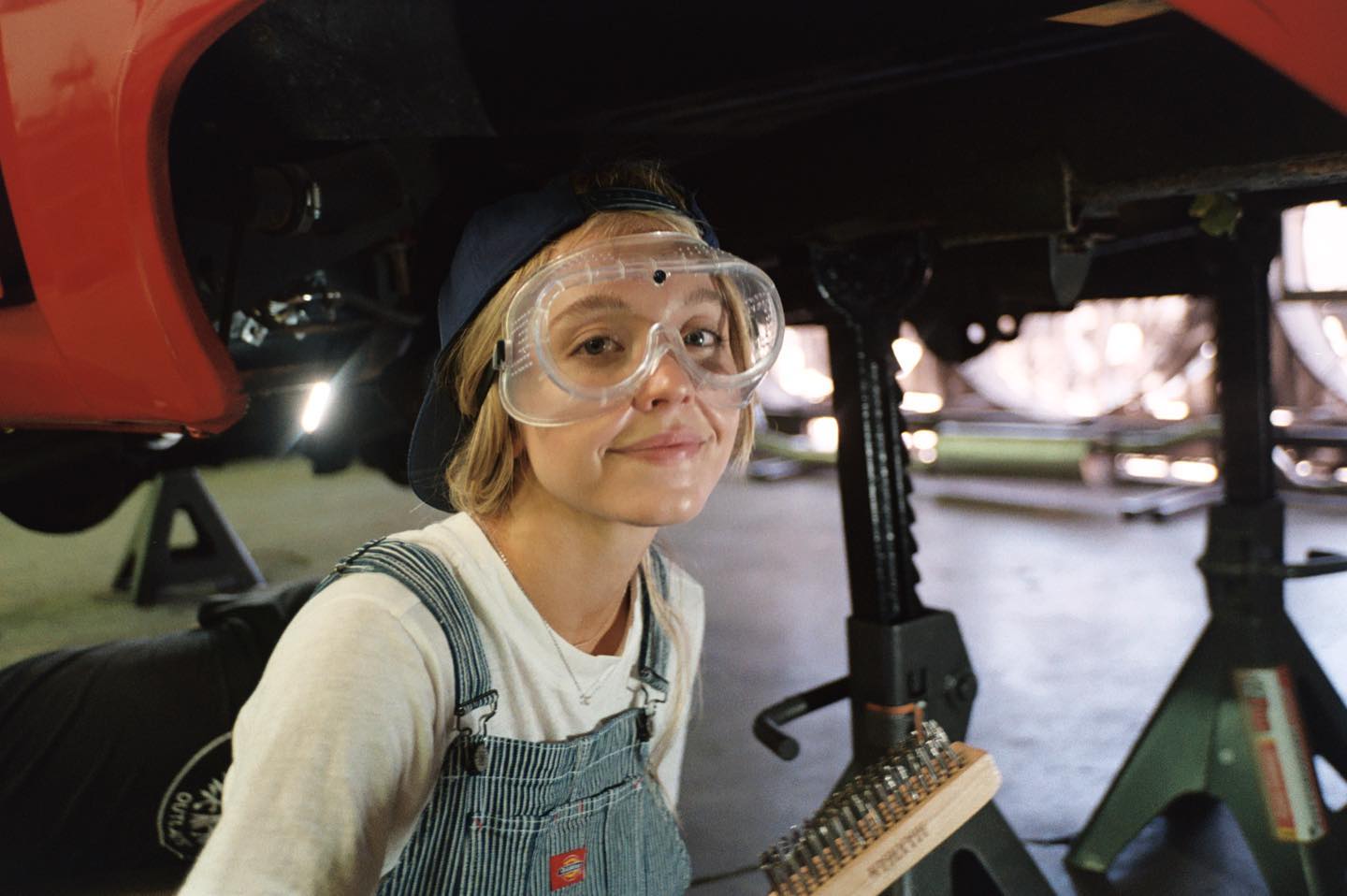 closeup portrait of a young woman, possibly a mechanic, working under a raised red car in a garage. she's wearing safety glasses, a denim overall, and a baseball cap. she appears to be using a wire brush on the vehicle's undercarriage. the overall setting is a garage or auto repair shop. the image has a candid and natural feel.